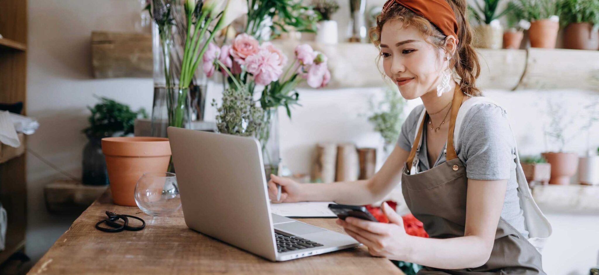 Woman in an apron working on a laptop and holding a smartphone in a floral workspace.