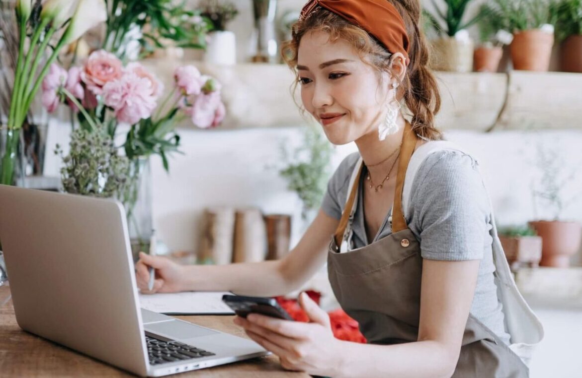 Woman in an apron working on a laptop and holding a smartphone in a floral workspace.